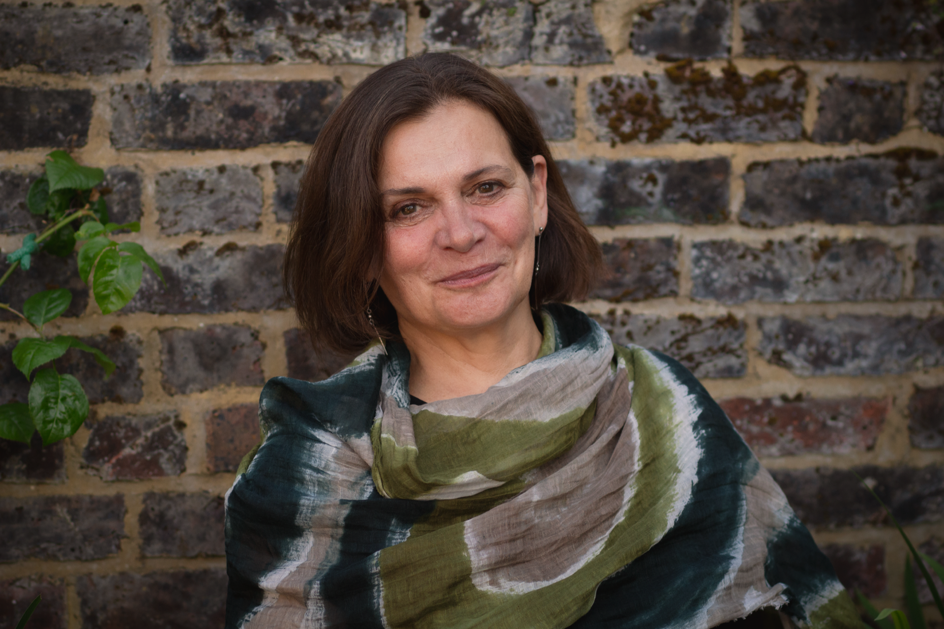 A woman with shoulder-length hair smiles at the camera, wearing a multi-colored scarf. She is standing in front of a textured brick wall with greenery in the background.